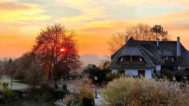 Une maison traditionnelle à toit de chaume, avec une cheminée bien visible, entourée d’arbres et sous un ciel dégagé.