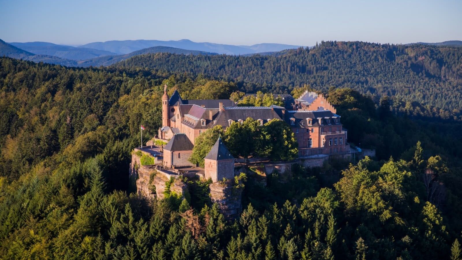 Una estructura que recuerda a un castillo, con varias edificaciones, ubicada en un acantilado rodeado de un bosque frondoso.