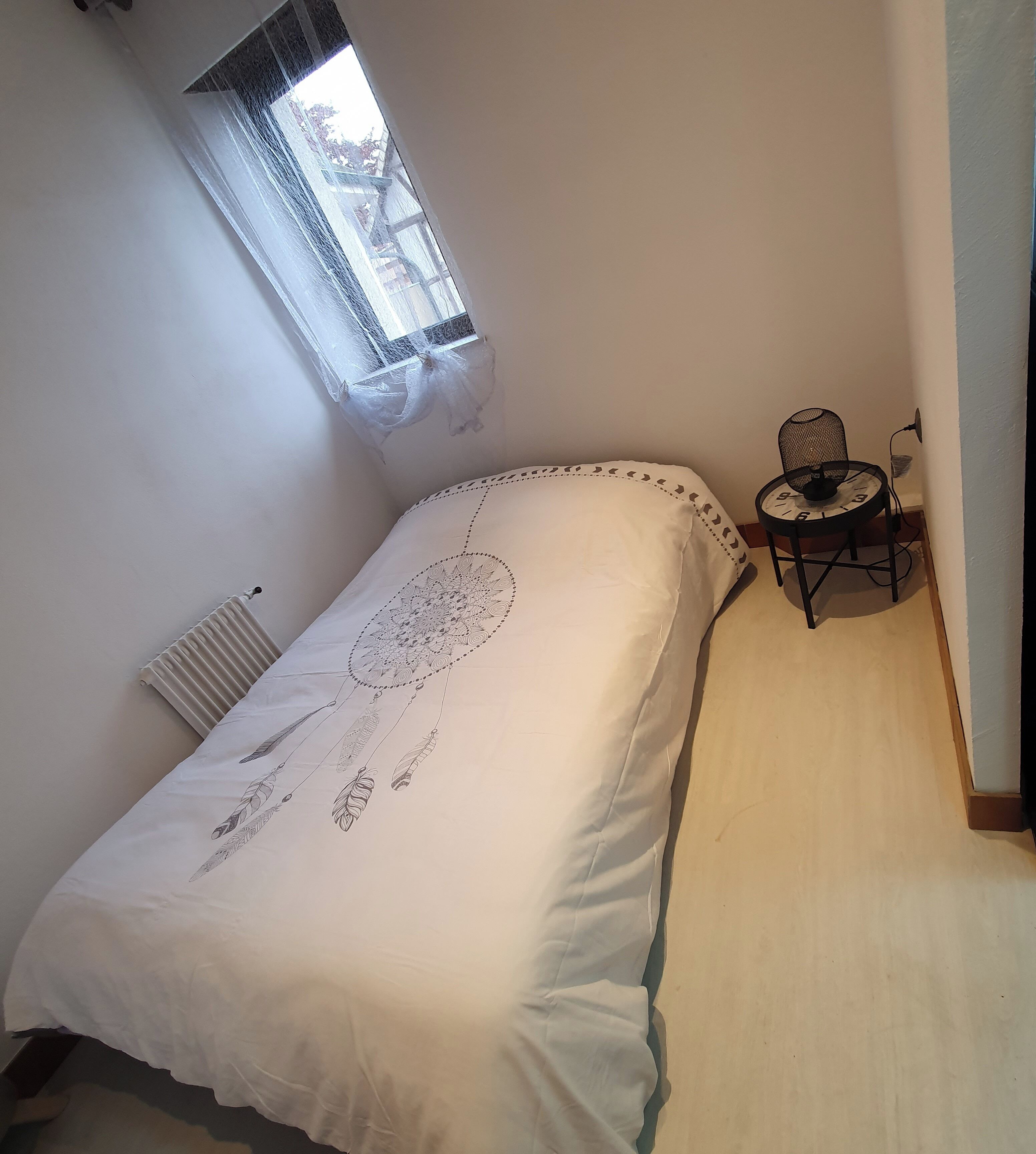 A bedroom with a bed, a radiator, a window, and a small table with a lamp.