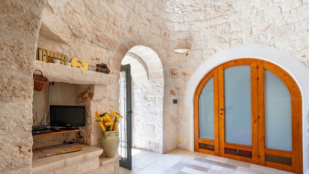 A stone-walled room with a television, a fireplace, and a large arched door. - Trullo Giuseppe by Rentbeat (Ostuni)