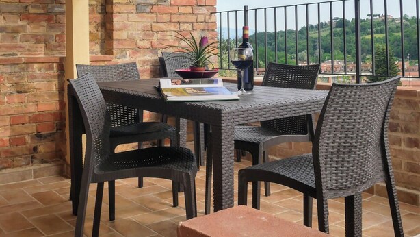 Um pátio externo com parede de tijolos, mesa com cadeiras, planta em vaso e taça de vinho.