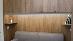 Quarto com cabeceira de madeira, sofá cinza e duas pequenas prateleiras com livros e difusor de vime.