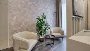 A modern hotel lobby with a geometric-patterned wall, two beige armchairs, a round table with a cup, and a large screen displaying content.