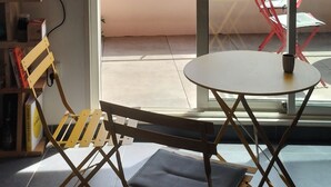 A room with a sliding glass door leading to a balcony. Inside, there is a small round table with a chair, a folding chair, and a bookshelf with books and a lamp.