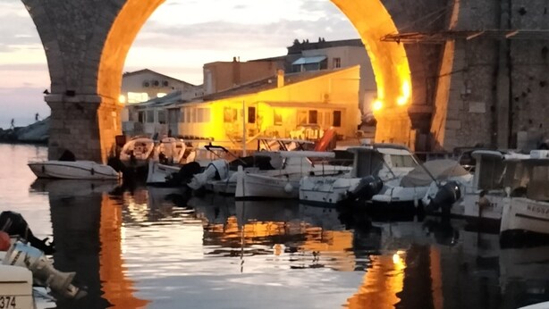 A stone bridge with an archway reflecting lights, boats docked in calm water, and a building with warm lighting in the background.
