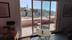A room with a large sliding glass door leading to an outdoor terrace. The room has a small round table and a chair, and there are framed pictures on the walls.