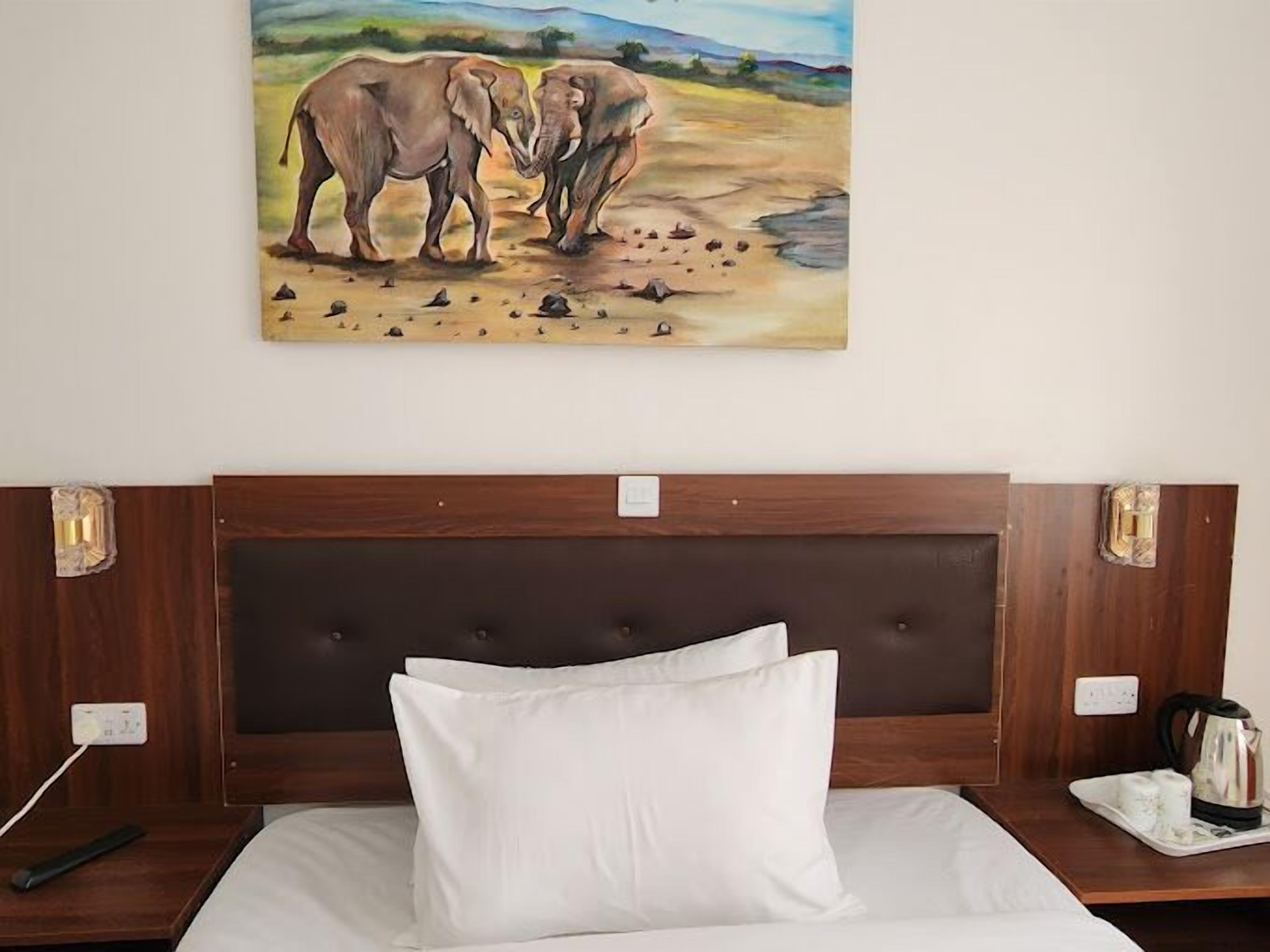 Habitación de hotel con un cuadro grande de elefantes en la pared, cabecero de madera, almohada blanca y una bandeja con hervidor y pañuelos.