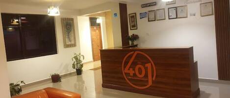 Un área de recepción con un escritorio de madera que muestra el número 401, un cartel de recepción, certificados enmarcados en la pared y plantas en macetas.