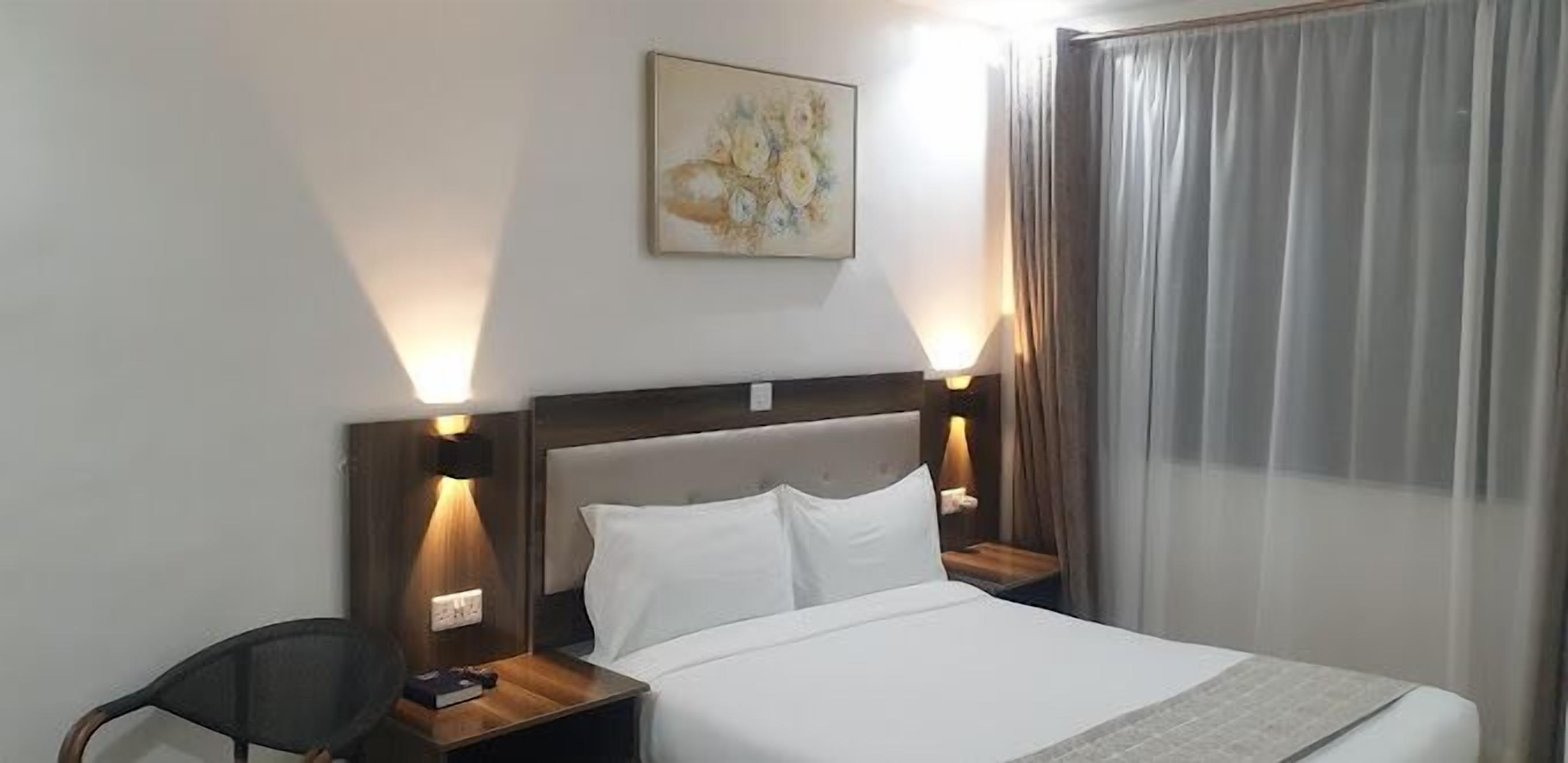 Een hotelkamer met een bed, twee bedlampen, een stoel, een schilderij aan de muur en een raam met gordijnen.
