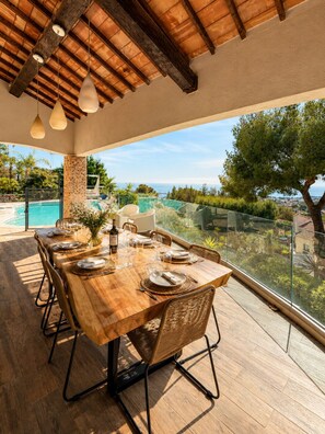 Un coin repas avec une table en bois, des chaises en osier et une vue sur une piscine et les environs.