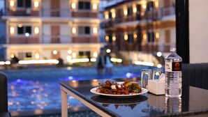 A dining table with a plate of food, a bottle of water, and glasses, overlooking a swimming pool and hotel buildings at dusk. - Shikali Resort (Khokana)