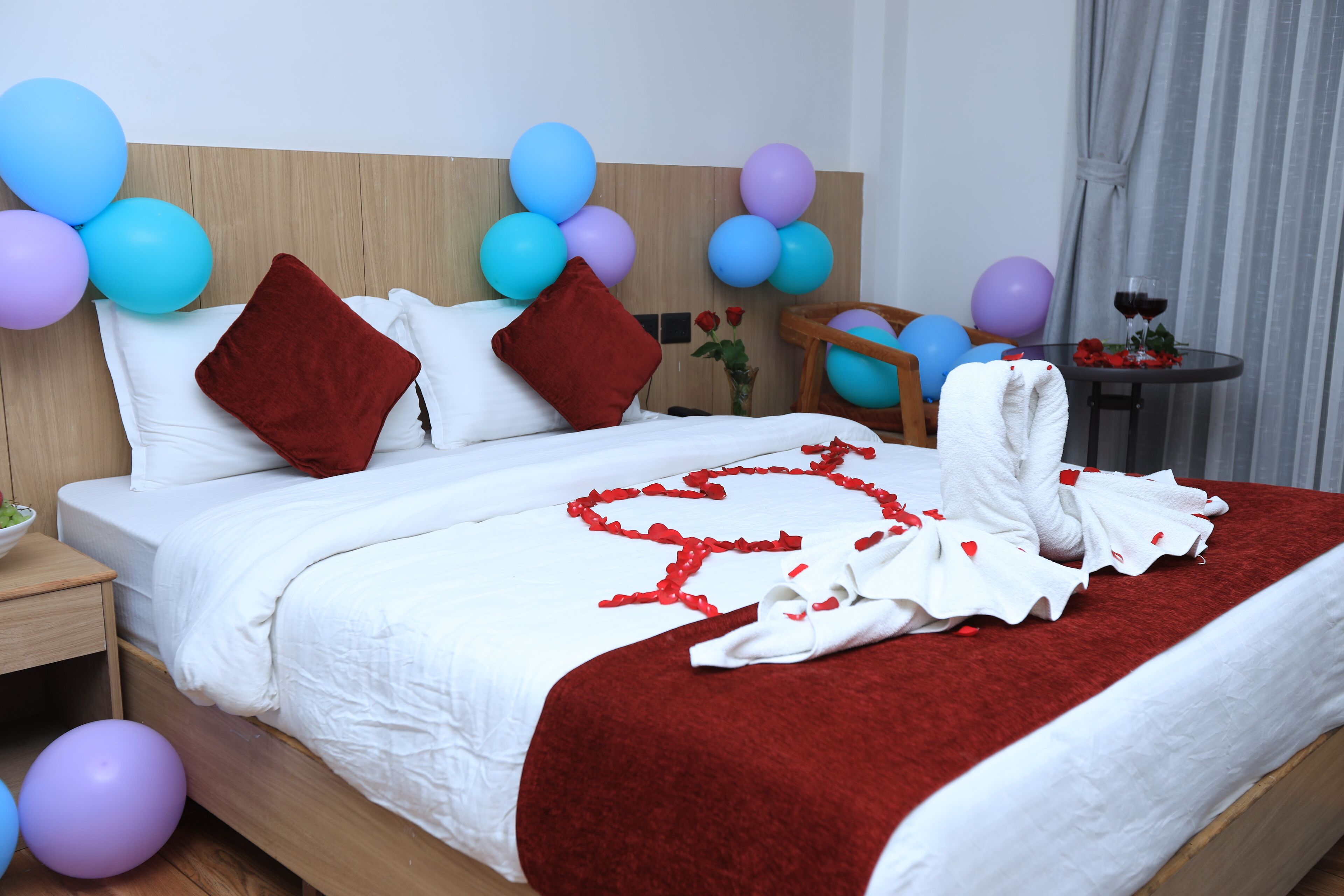 A hotel room with a neatly made bed, a wooden headboard, and a small table with a glass of red wine and roses.