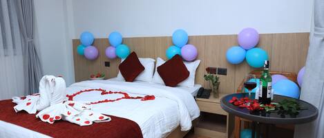Een hotelkamer met een net opgemaakt bed, een klein tafeltje met een fles en glas, en een muur versierd met ballonnen.