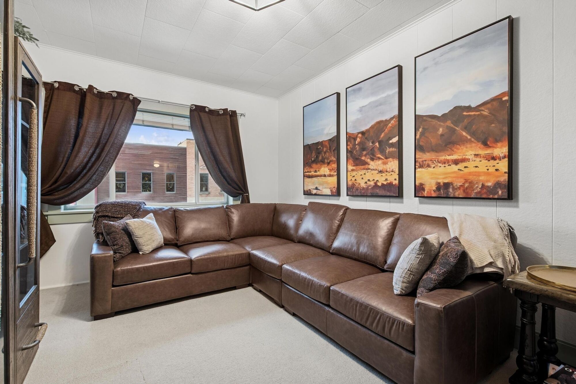 A living room with a brown leather sectional sofa, a small round table, and three framed landscape paintings on the wall.