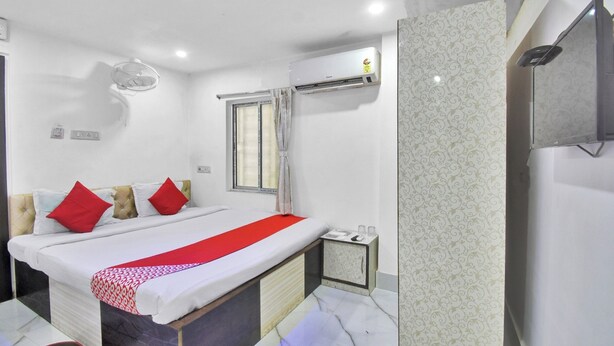 A hotel room with a bed, a window with curtains, an air conditioning unit, a small bedside table, and a wall-mounted TV. - Hotel Palace- Near Ruby Hospital (Kolkata)