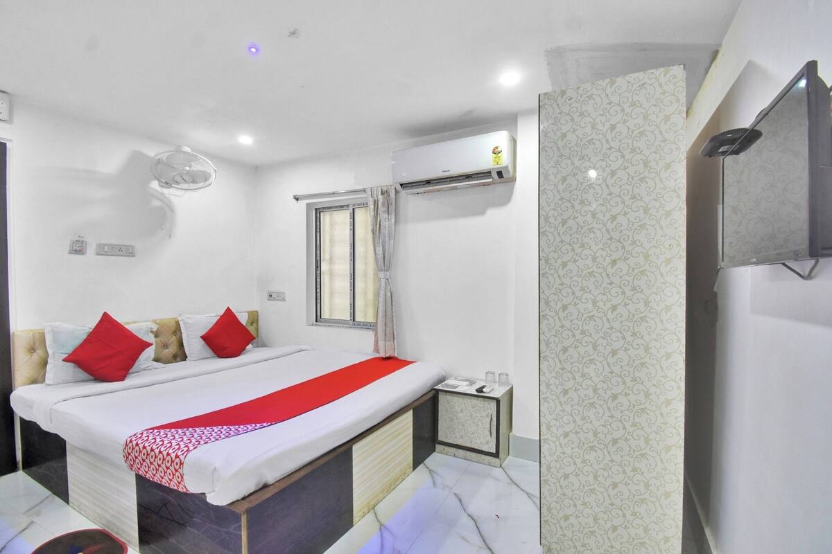 A hotel room with a bed, a window with curtains, an air conditioning unit, a small bedside table, and a wall-mounted TV.
