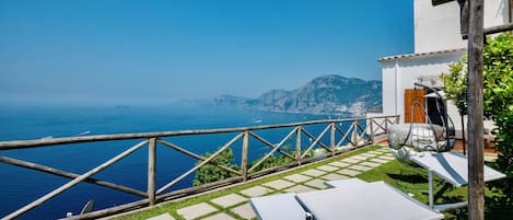 A terrace with a view of the sea, a wooden fence, and a white building with a door and a chair.