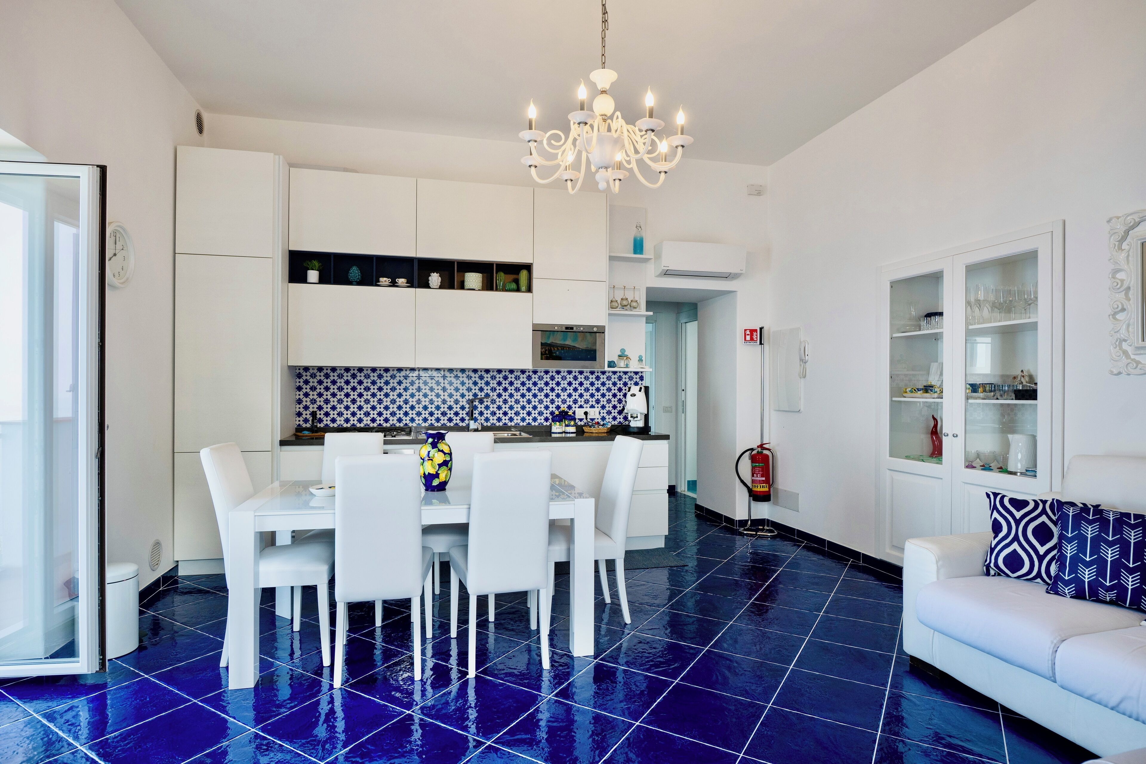A modern kitchen with white cabinets, a blue tiled backsplash, and a dining area with white chairs.