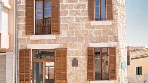 Un edificio de piedra de dos pisos, con contraventanas de madera, una entrada visible en la planta baja y un cielo despejado.