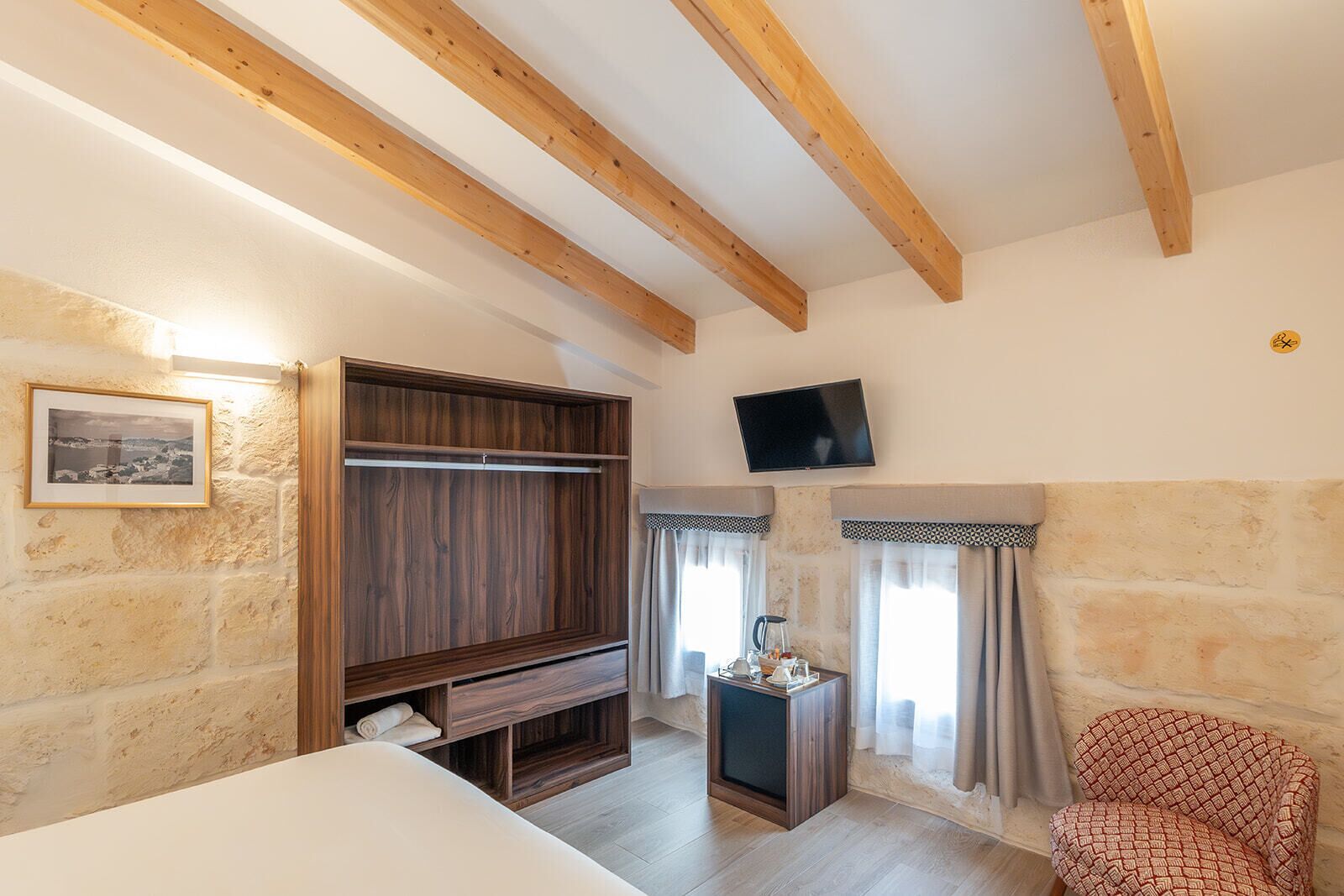 A room with exposed wooden beams, a wooden wardrobe, a television, a small table with a kettle, and a patterned chair.