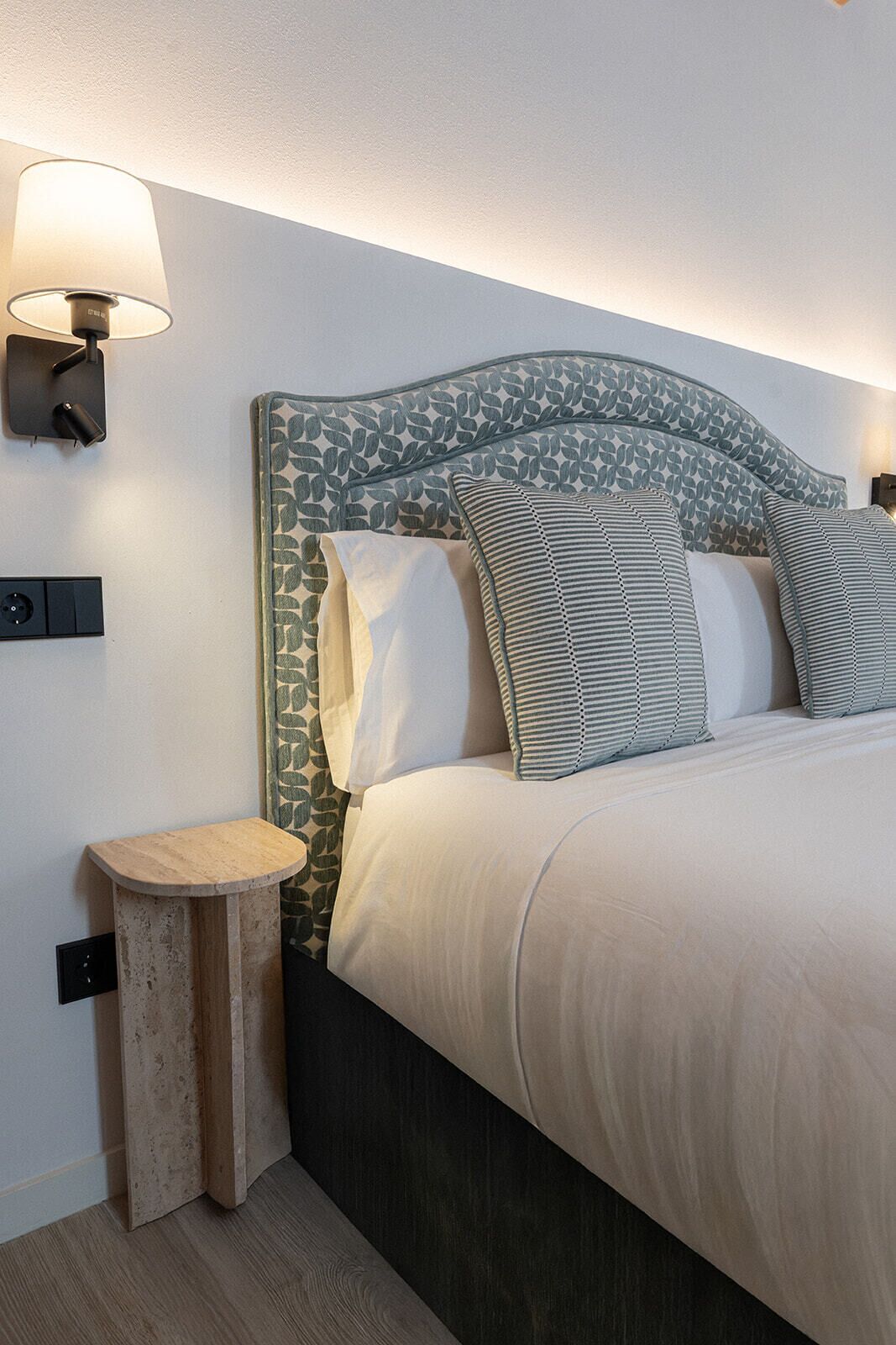 A neatly made bed with patterned pillows, a headboard with a leaf design, a bedside lamp, and a small wooden nightstand.