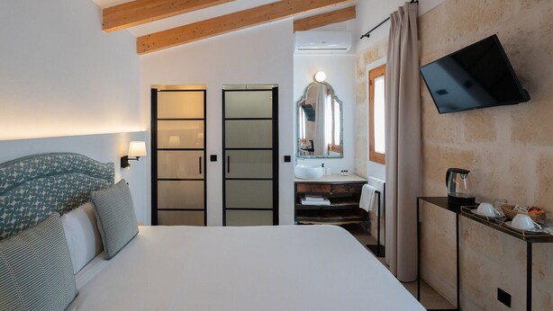 Habitación de hotel con cama, televisor, baño con espejo y vigas de madera en el techo.