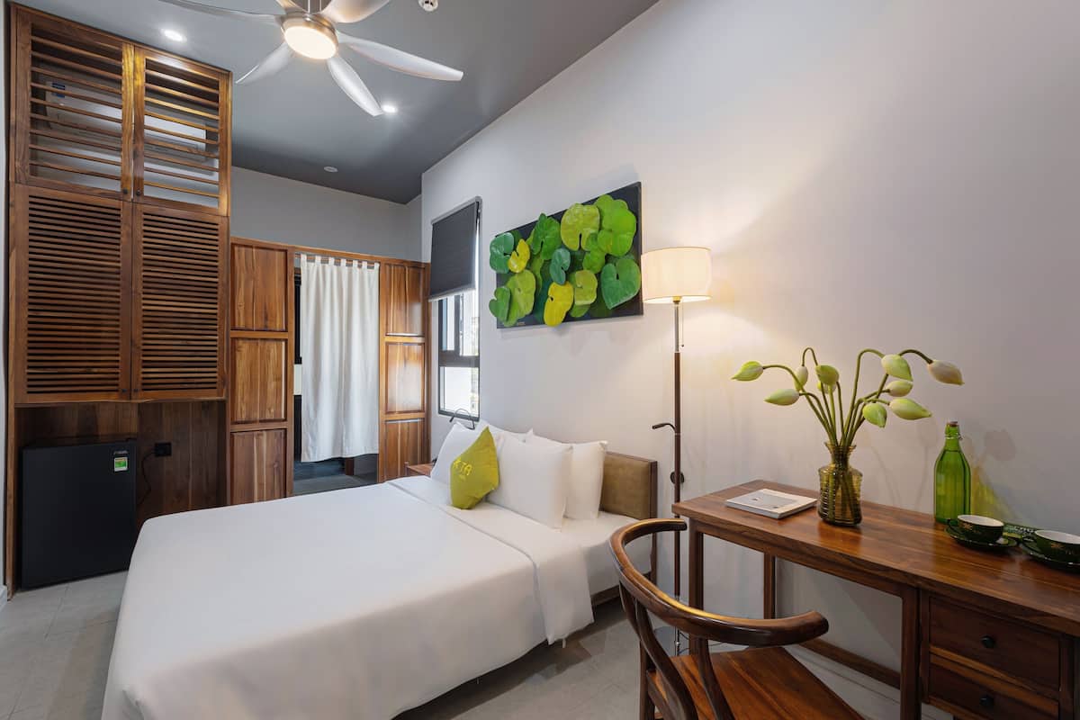 Une chambre d’hôtel comprenant un lit, un bureau, une chaise, une lampe, une petite table, un vase avec des fleurs, un tableau au mur et un ventilateur de plafond.