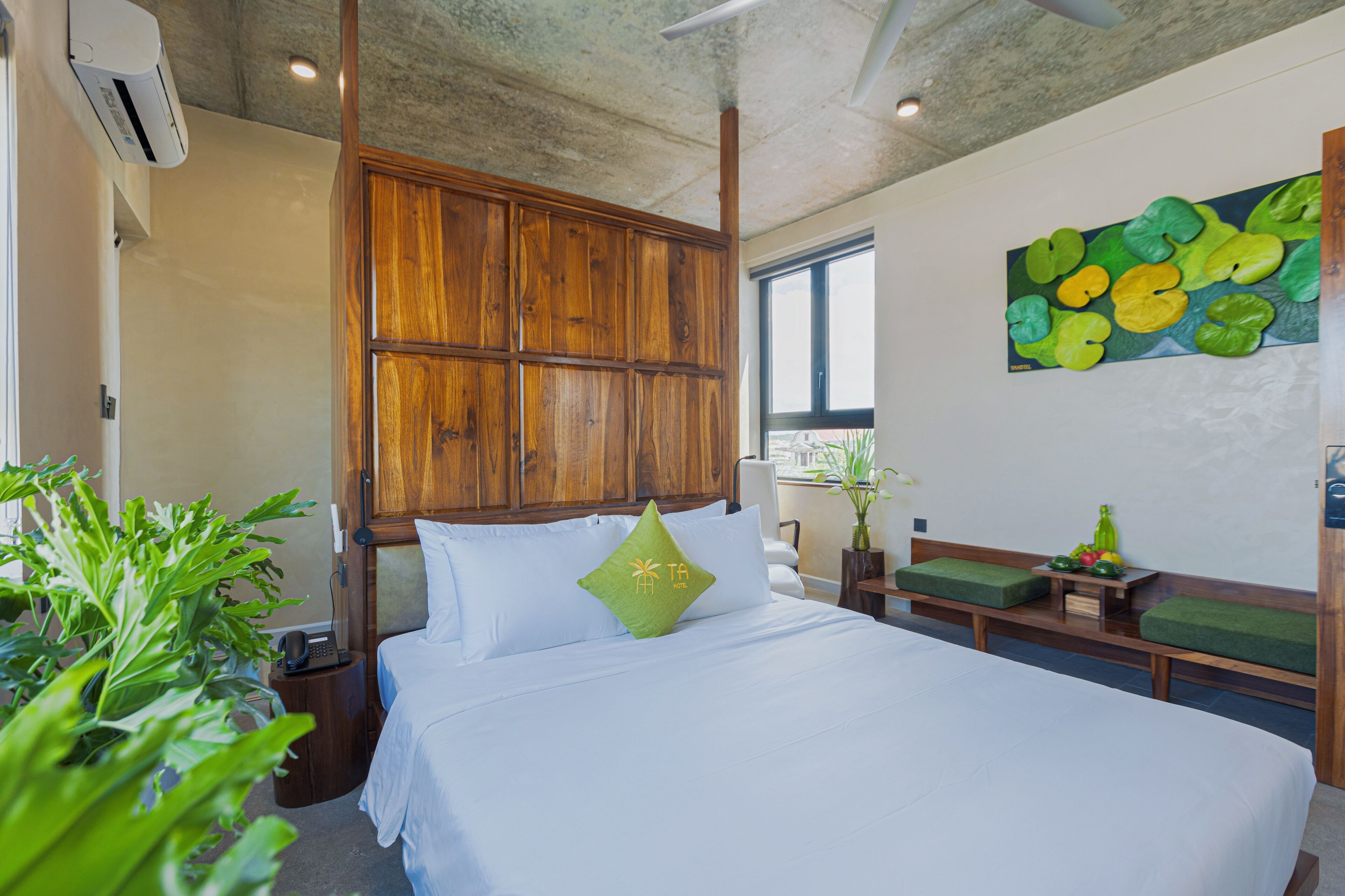 A hotel room with a large bed, wooden headboard, green cushions, a small table with fruit, and a painting of lily pads on the wall.