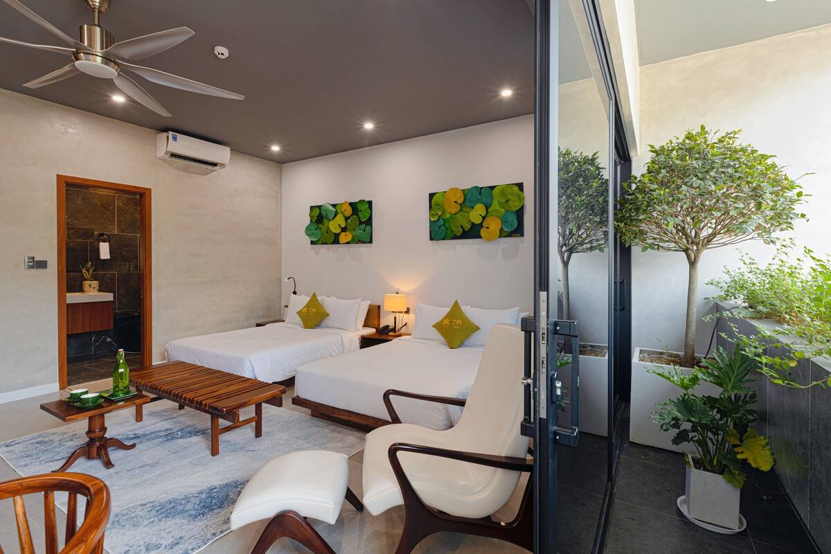 Une chambre d’hôtel avec deux lits, un ventilateur de plafond, un appareil de climatisation, une table en bois, un canapé, une chaise et une plante en pot.