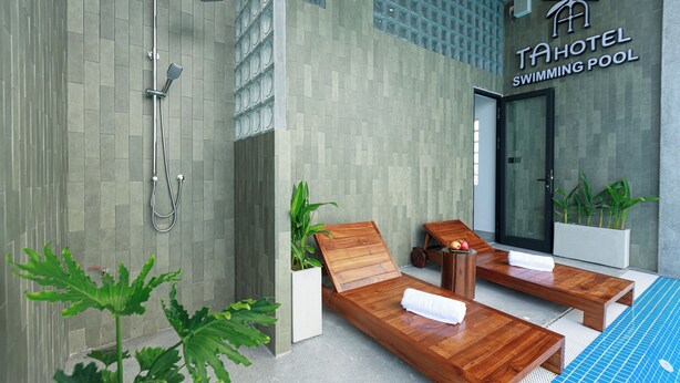 A modern hotel swimming pool area with a shower, two wooden loungers, and a sign reading "TA HOTEL SWIMMING POOL".