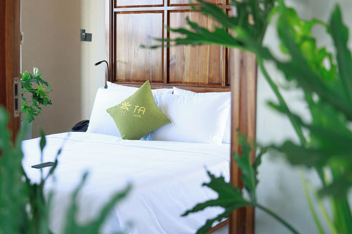 Un lit bien fait, avec du linge de lit blanc et un seul coussin vert, une tête de lit en bois et une plante sur le côté gauche.