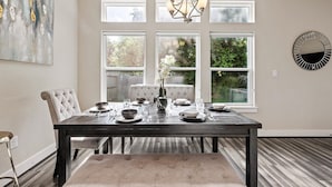 Comedor con mesa de madera oscura dispuesta para cuatro comensales, una butaca tapizada, una araña y un ventanal con vistas a follaje.
