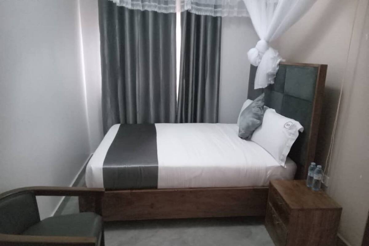 A bedroom with a bed, a bedside table with bottles, a chair, and a window with curtains.
