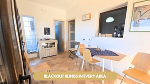 A room with blackout blinds in every area, a dining table with chairs, a kitchen area, and a large window. - Dammuso - Cala Cinque Denti - AFFITTI\'CA (Pantelleria)
