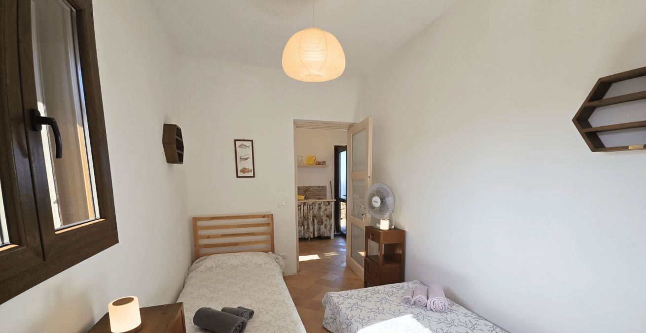 Una habitación pequeña y acogedora con dos camas, una mesita de noche con lámpara y un ventilador. Indica que se proporcionan toallas y ropa de cama.