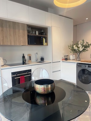 A modern kitchen with white cabinets, a microwave, a sink, and a dishwasher. There is a glass table with a decorative bowl. - The Rubik Executive Apartment (Cape Town)
