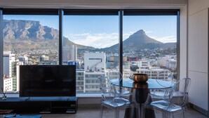 Una habitación moderna con un ventanal grande que ofrece vistas a la ciudad, incluyendo montañas, un comedor con una mesa y sillas de vidrio, y un televisor de pantalla plana sobre un mueble.