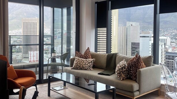 Una sala de estar moderna con sofá, mesa de centro y una mesa de comedor de vidrio. Grandes ventanales ofrecen vistas a la ciudad.