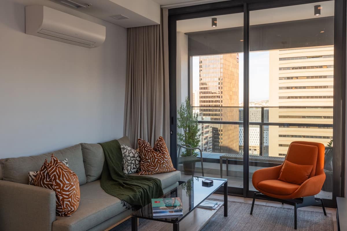 Uma sala de estar moderna com sofá, poltrona e mesa de centro, grandes janelas com vista para a cidade e um vaso com planta.