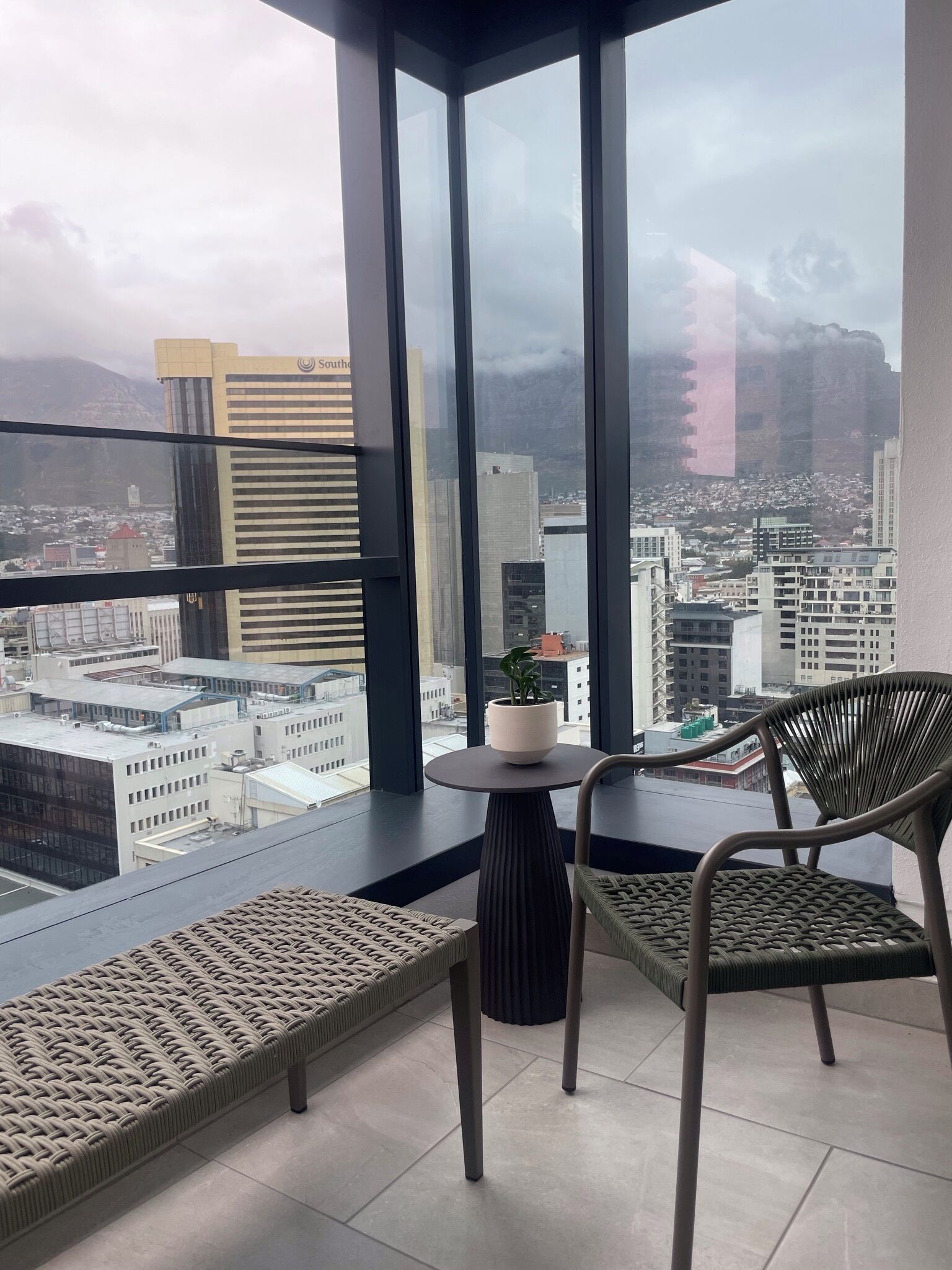 A balcony with a view of a cityscape, featuring modern buildings and a mountain in the background. There is a small round table with a potted plant, and two wicker chairs.
