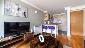 Un salon moderne avec un téléviseur à écran plat, un meuble de rangement en bois et une table basse ronde. On aperçoit à l’arrière-plan une cuisine avec des tabourets de bar et un coin repas.