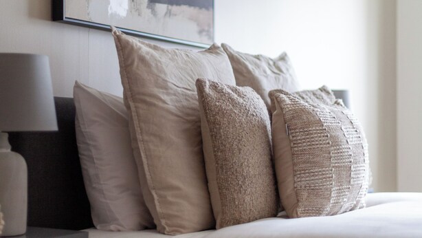 Un lit bien fait, avec une literie et des oreillers de couleur beige, une table de chevet avec une lampe, et un grand tableau abstrait accroché au mur.