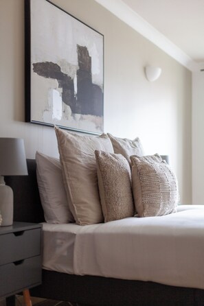 Een opgemaakt bed met beige beddengoed en kussens, een nachtkastje met een lamp, en een groot abstract schilderij aan de muur.