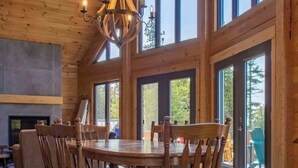A wooden interior with a large dining table, chairs, a chandelier, and large windows.