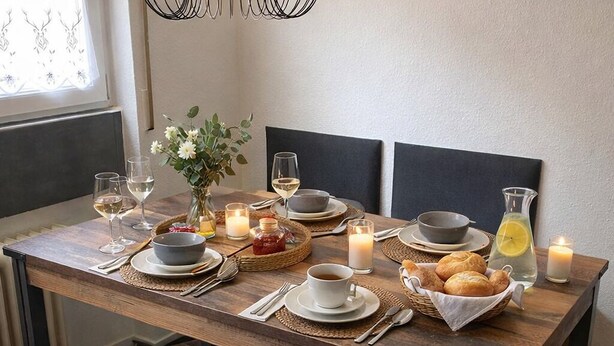 Una mesa de comedor dispuesta con platos, vasos y velas, con un banco y almohadones al frente.