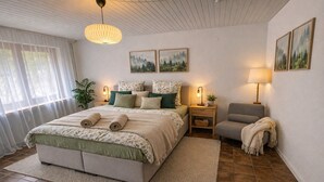 Un dormitorio con cama, mesitas de noche, una silla, una lámpara y dos cuadros en la pared.