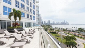 A modern hotel with a rooftop lounge area, a city skyline view, and a waterfront promenade.