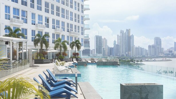 A rooftop pool with lounge chairs, palm trees, and a city skyline in the background.