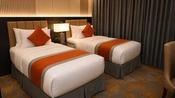 Habitación de hotel con dos camas, una mesita de noche con lámpara y vistas al exterior a través de cortinas.