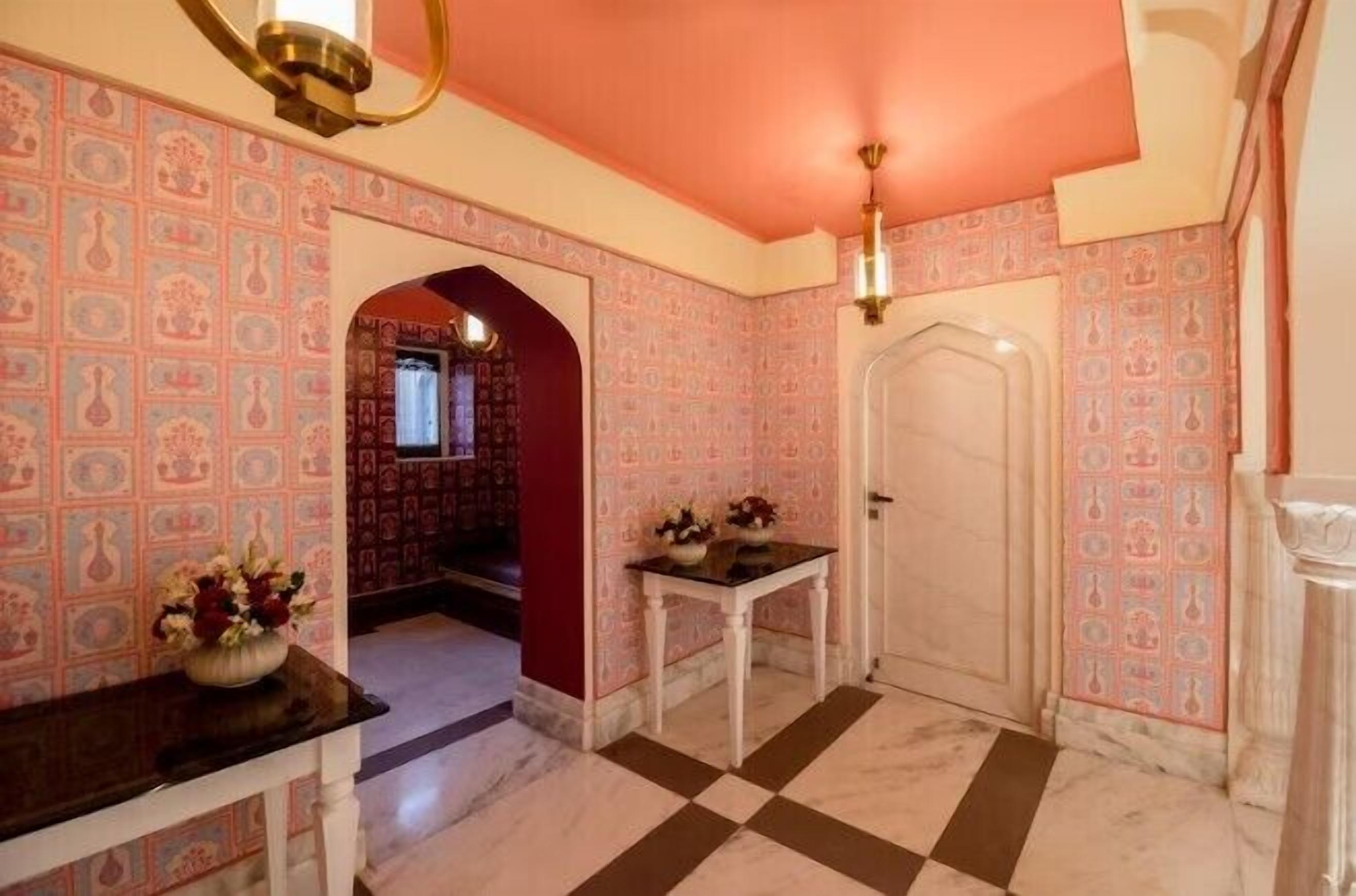 Un pasillo con papel tapiz estampado, suelo de cuadros, una puerta blanca y una pequeña mesa con flores.